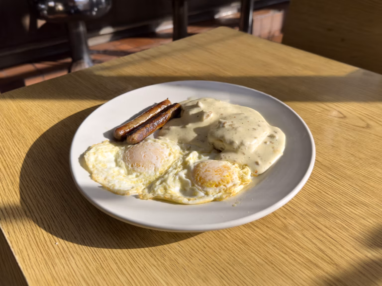 A comforting and hearty Northside Grill combo plate featuring two fluffy biscuits completely covered in rich, savory sausage gravy, served alongside two eggs any style and three savory sausage links on a rustic dining table.
